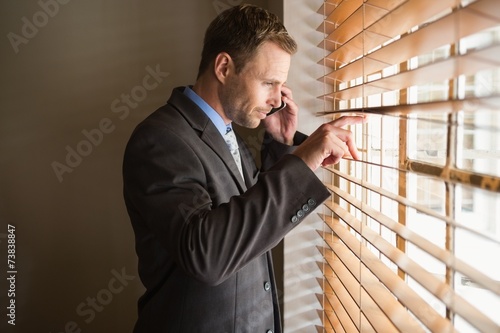 Businessman peeking through blinds while on call