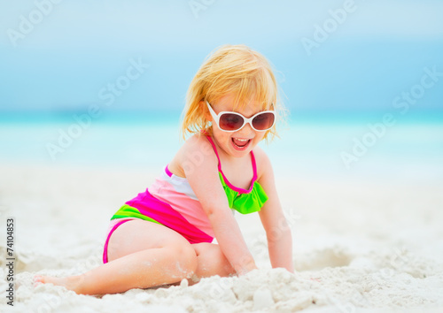 Smiling baby girl in sunglasses playing with sand on beach