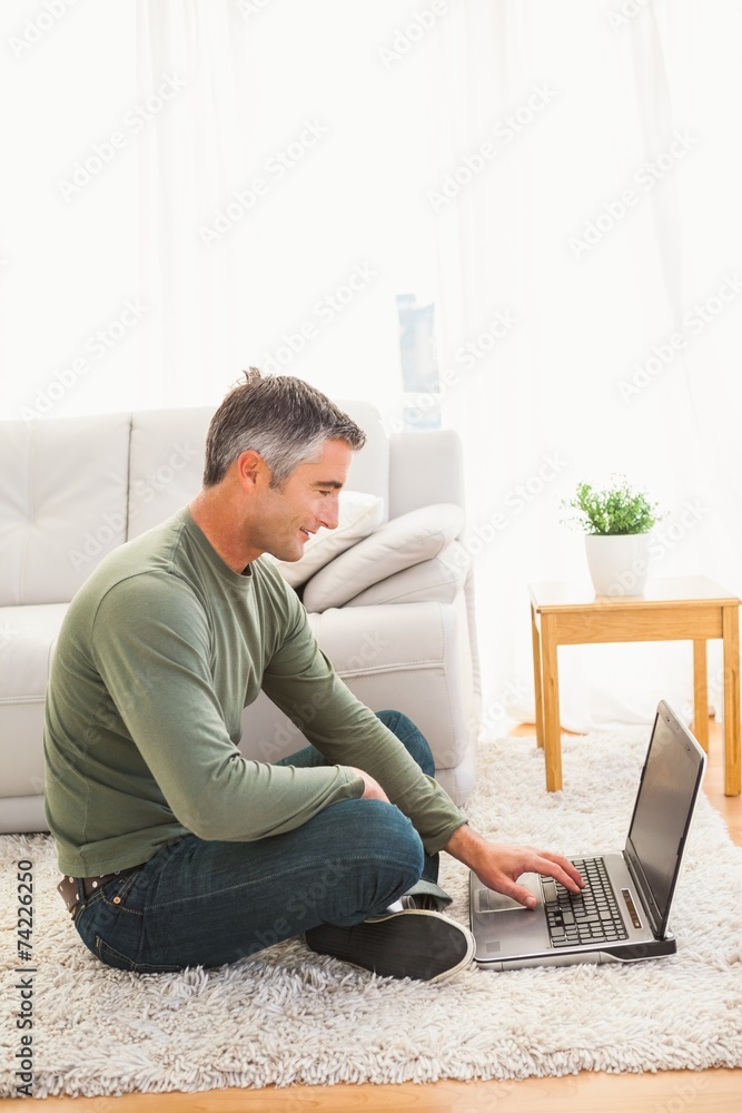 Smiling man sitting on carpet using laptop