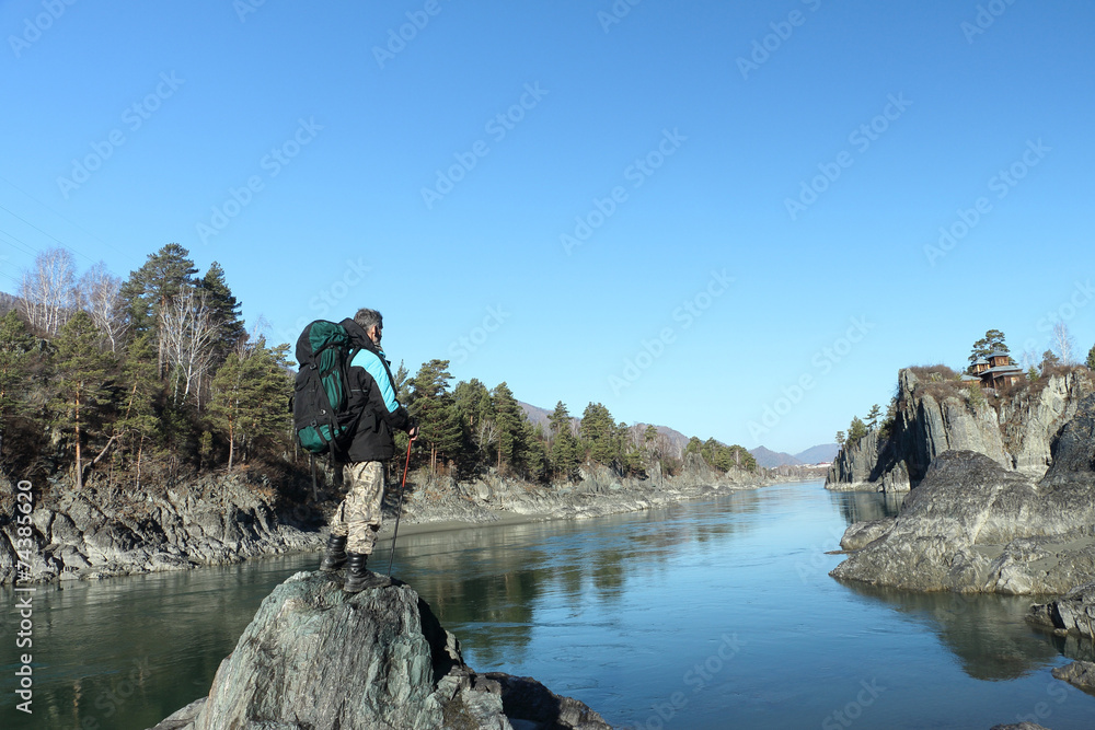 The traveler with a backpack standing on a stone on the river ba