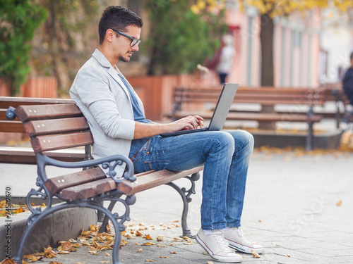 Bild auf Leinwand Young man sitting on the park bench with laptop on his lap