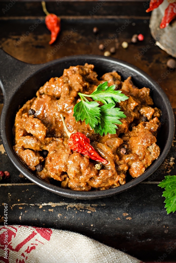 Stewed beef liver with tomatoes sauce in iron pot on iron dark background Stewed beef liver with tomatoes sauce in iron pot on iron dark background
