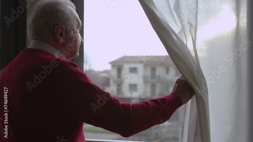 Worried elderly man moves the curtain and looks out the window