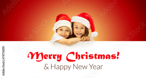 Happy little kids in Santa hat peeking from behind blank sign