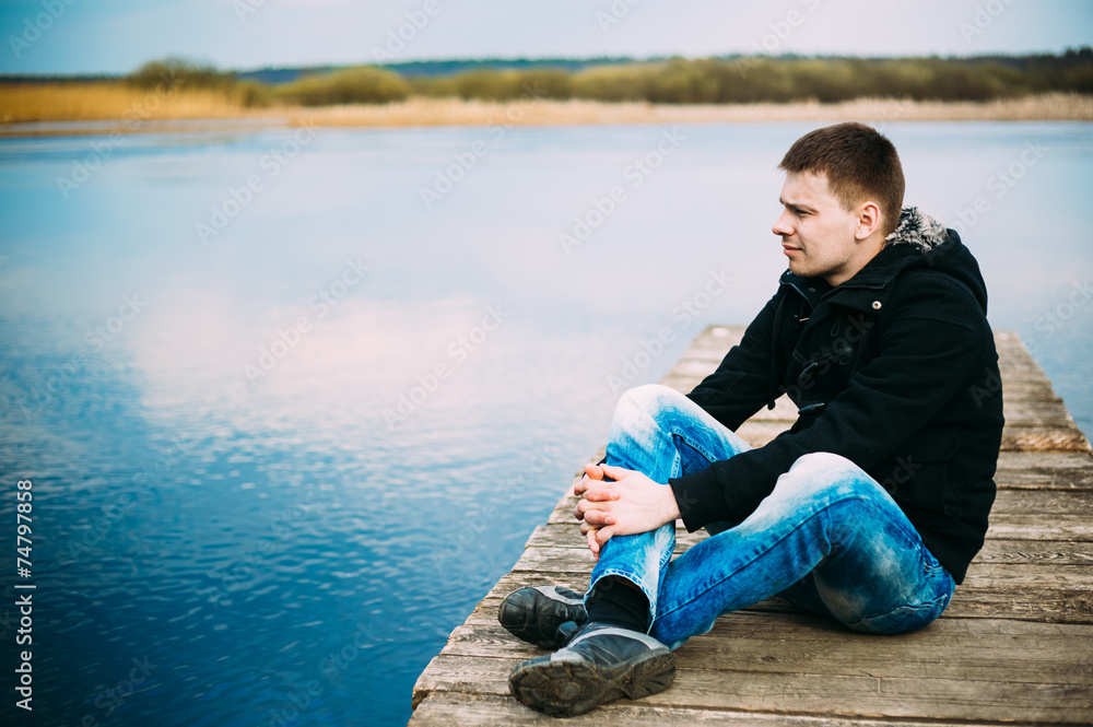 Young handsome man sitting on wooden pier, relaxing,  thinking,