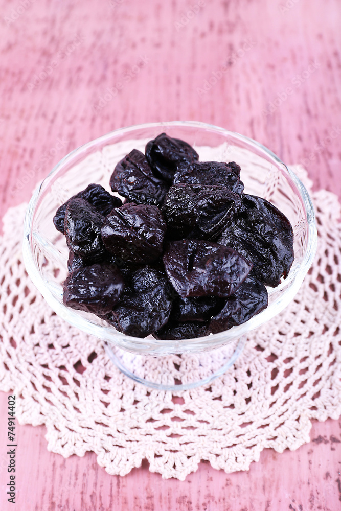 Prunes in glass bowl on lace doily on color wooden background Prunes in glass bowl on lace doily on color wooden background