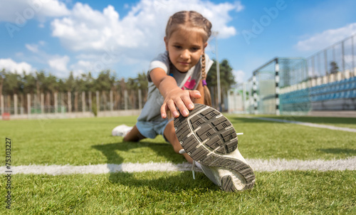 young girl stretching legs on soccer field at sunny day