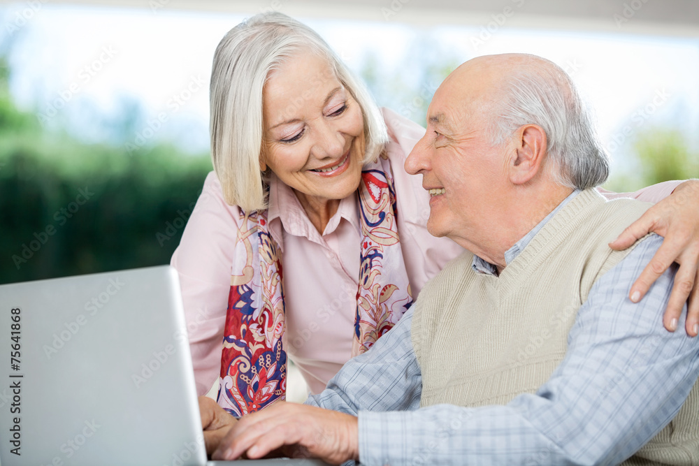 Smiling Senior Couple Looking At Each Other While Using Laptop