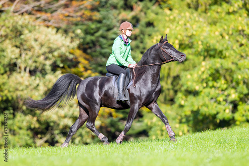 Young girl riding a black horse in autumn time near forest.