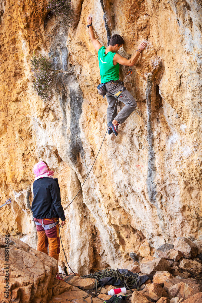 Young man lead climbing on cliff, female climber belaying