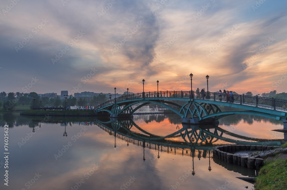 Fototapeta premium Пешеходный мост на пруду в парке летним вечером на фоне заката