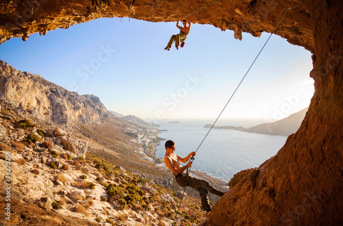 Rock climber climbing on roof in cave, his partner belaying