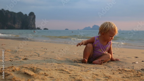 blonde baby girl in purple swimsuit play with seashell near sand