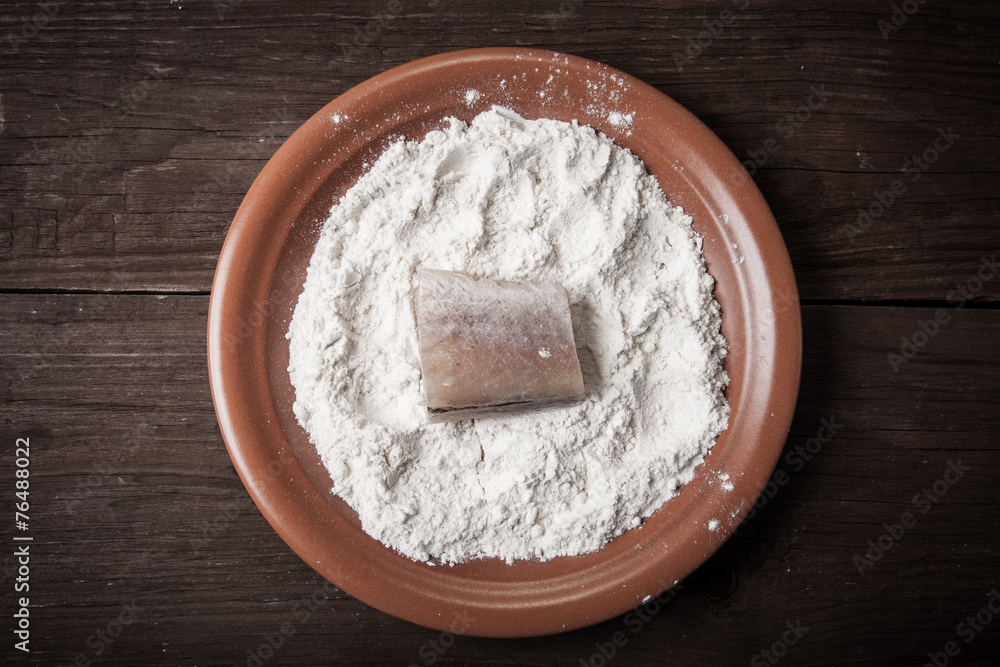 One piece of fresh fish with flour on a plate on a wooden backgr One piece of fresh fish with flour on a plate on a wooden backgr