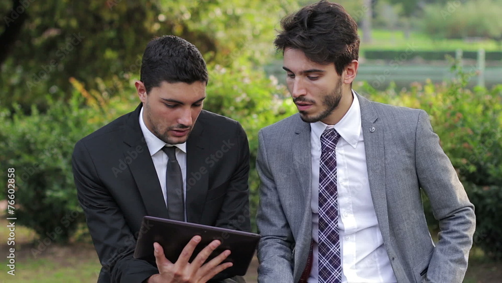 Two business people with tablet computer talk about a project on a park bench Two business people with tablet computer talk about a project on a park bench