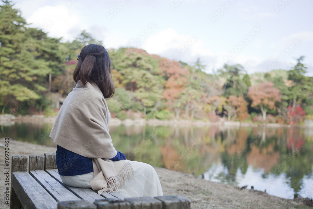 秋の紅葉の公園の池の前のベンチに座る女性