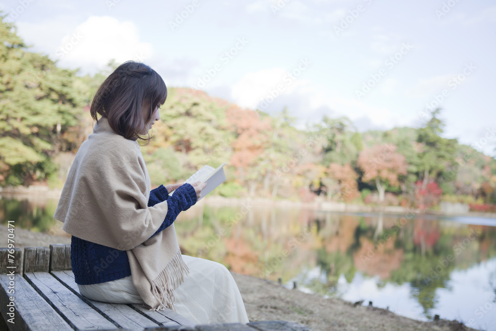 秋の紅葉の公園の池の前のベンチで本を読む女性