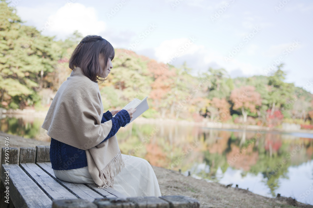 秋の紅葉の公園の池の前のベンチで本を読む女性