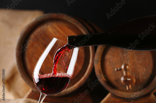 Pouring red wine from bottle into glass with wooden wine casks