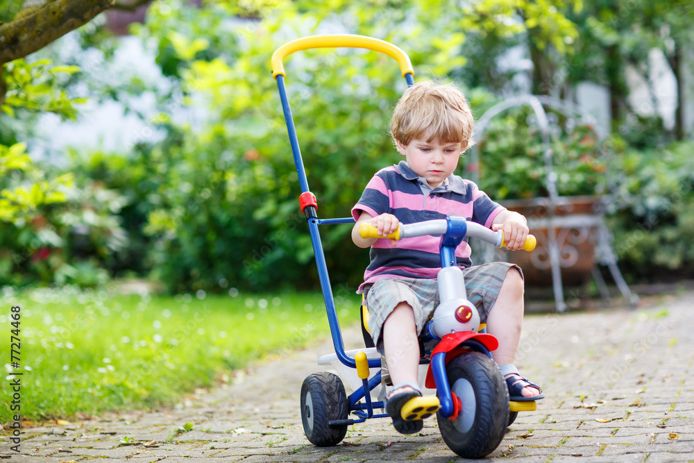 Active blond kid boy driving tricycle or bicycle in domestic gar Active blond kid boy driving tricycle or bicycle in domestic gar