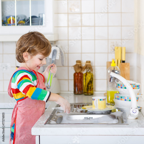 Happy little blond kid boy washing dishes in domestic kitchen