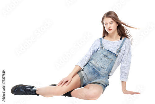 Young fashion girl in jeans overalls sitting isolated