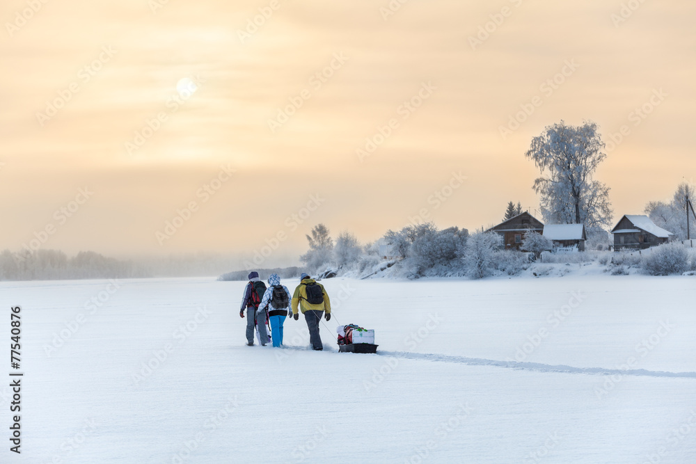 Winter travelers hiking on lake ice at sunset over village