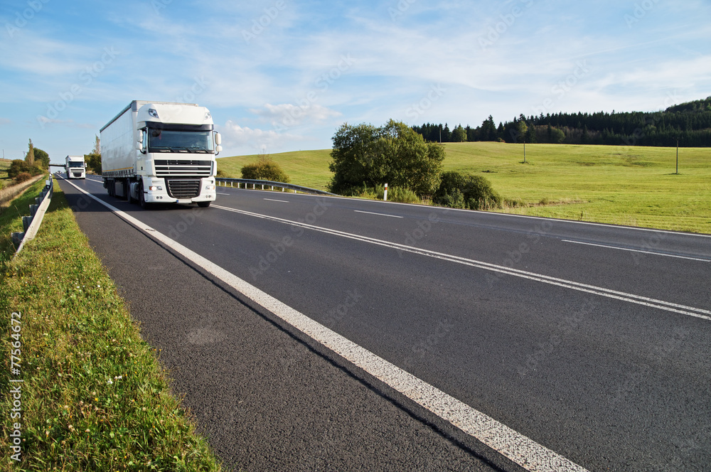Asphalt road in a rural landscape, the arriving two white trucks