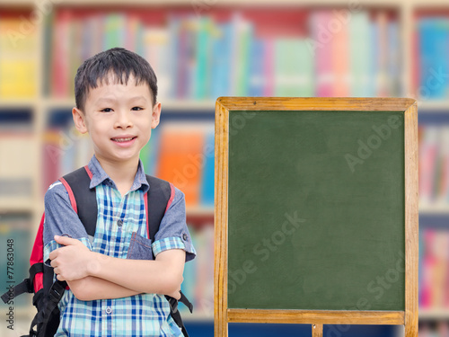 Happy Asian boy standing near by empty chalkboard in library photo