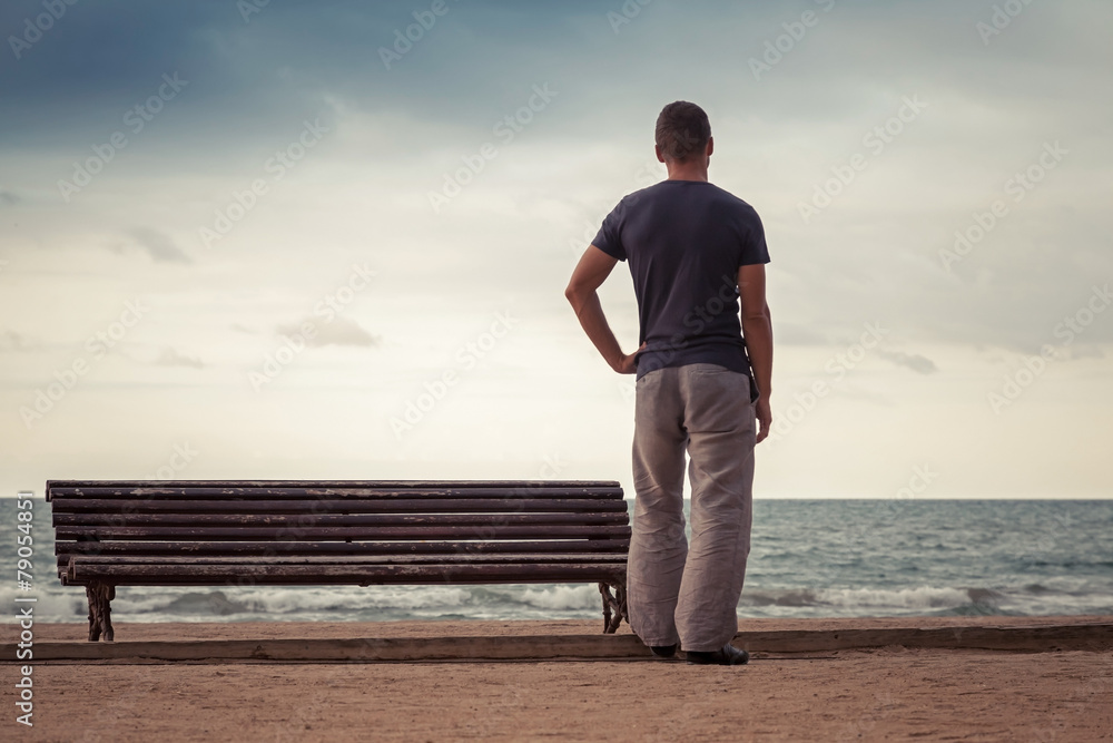 Young man stands near old bench on the sea coast