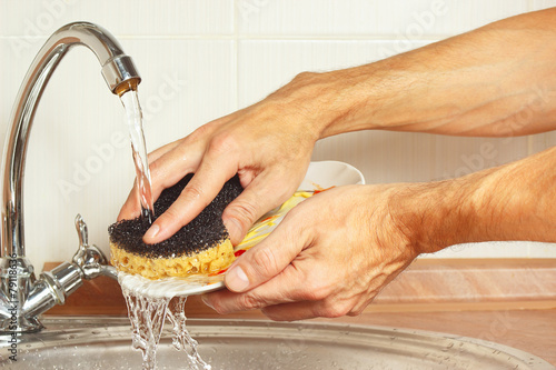Hands wash the dirty dishes under running water in the kitchen