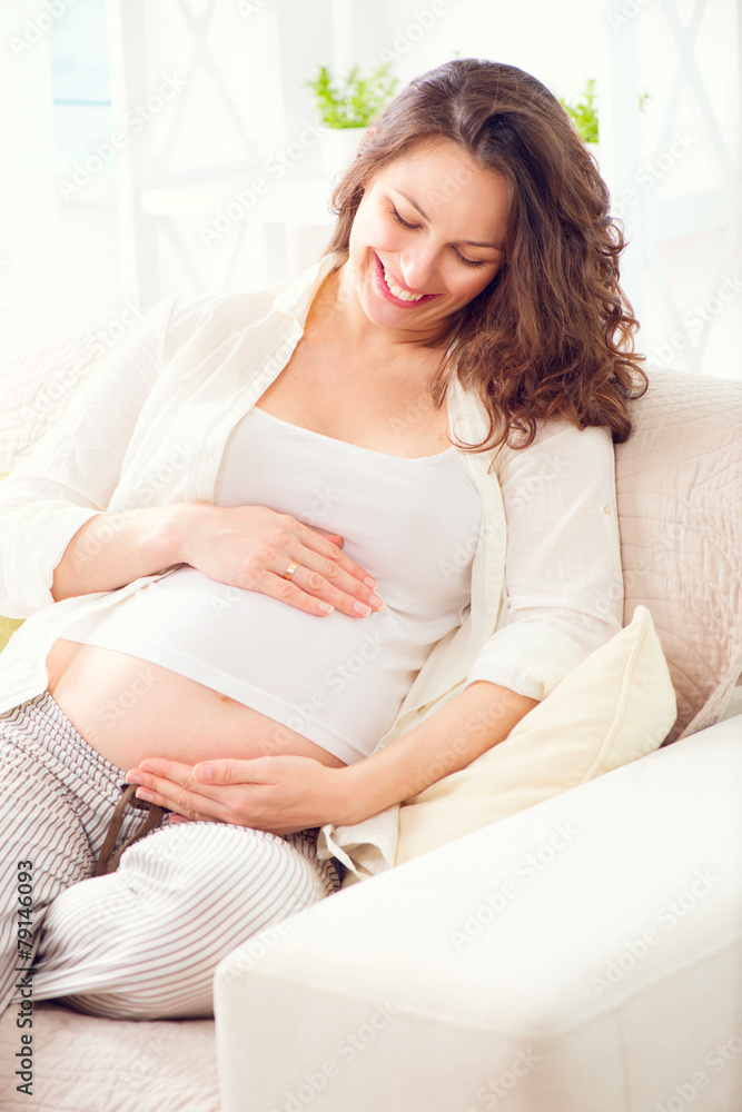 Pregnant happy woman sitting on a sofa and caressing her belly Pregnant happy woman sitting on a sofa and caressing her belly