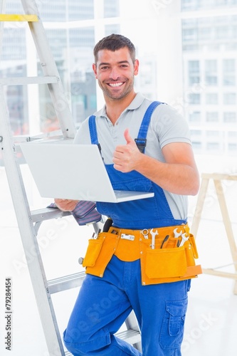 Smiling handyman with laptop gesturing thumbs up in office