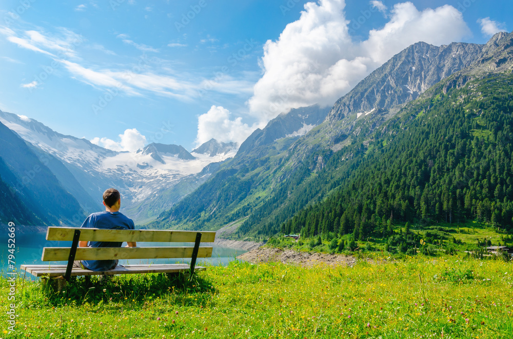 A young man sits on bench beside an azure mountain lake