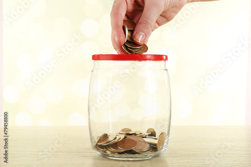 Female hand putting coins in glass bottle on bright background