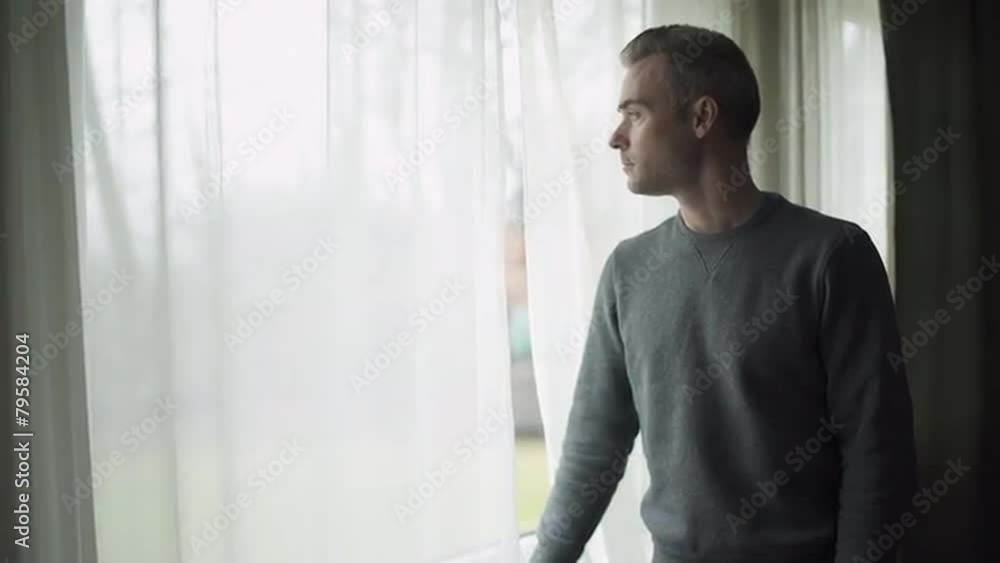 A depressed man stands at a window on a stormy cold day and looks out the window. A depressed man stands at a window on a stormy cold day and looks out the window.