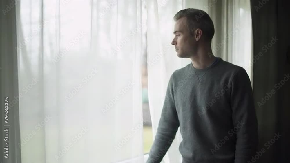 A depressed man stands at a window on a stormy cold day and looks out the window. A depressed man stands at a window on a stormy cold day and looks out the window.