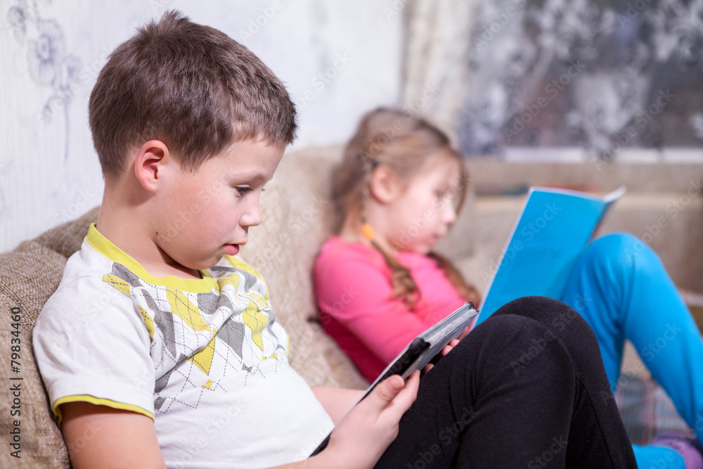 Brother sits with electronic tablet in hands, sister reads book Brother sits with electronic tablet in hands, sister reads book