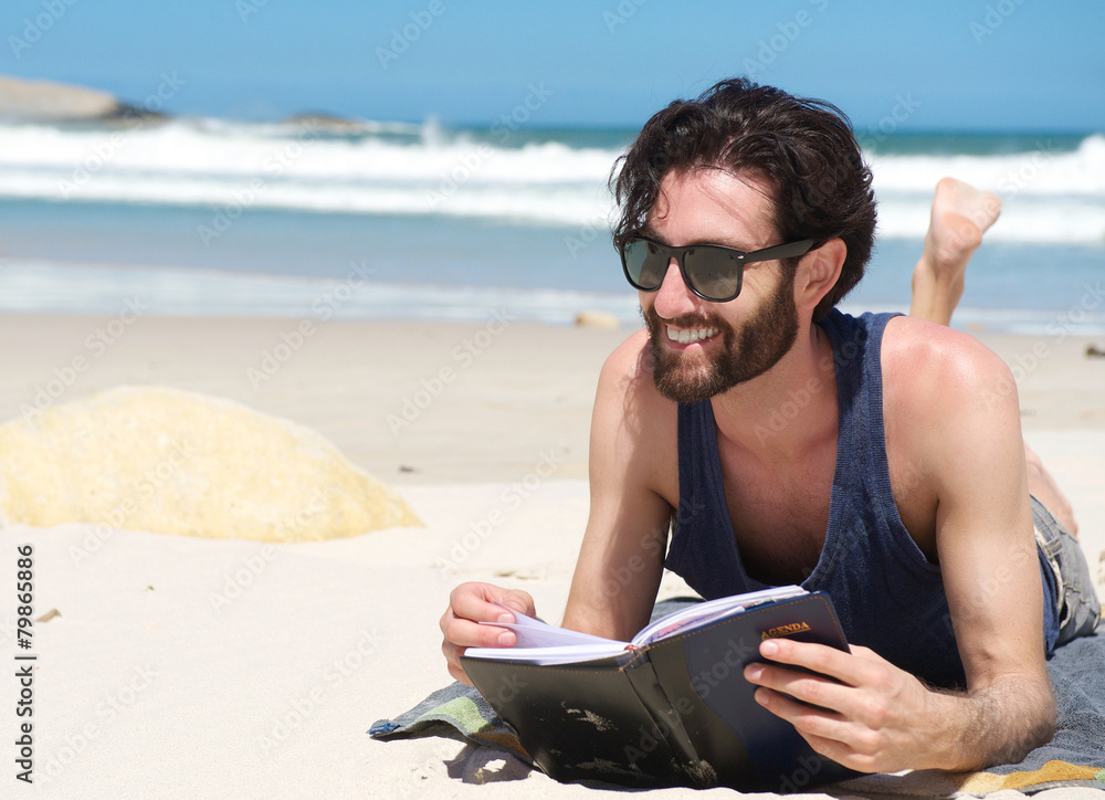 Handsome young man smiling and reading book on the beach
