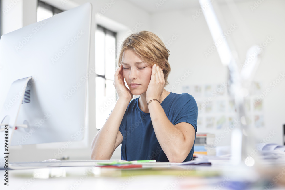 Portrait of young woman with headache at her desk in a creative office Portrait of young woman with headache at her desk in a creative office