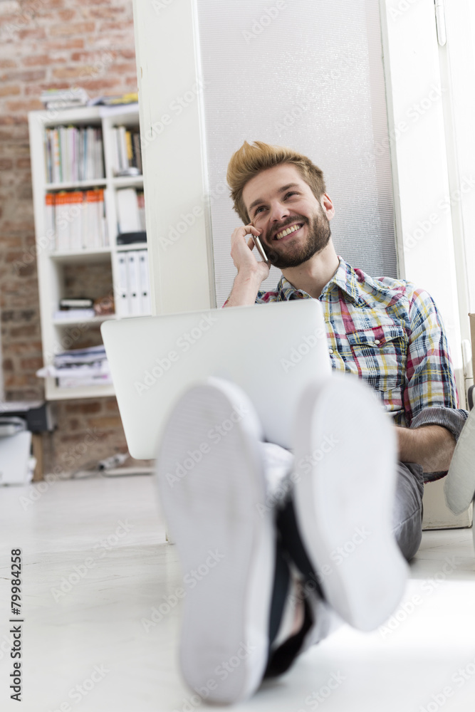 Young man sitting on floor in office using laptop and cell phone Young man sitting on floor in office using laptop and cell phone