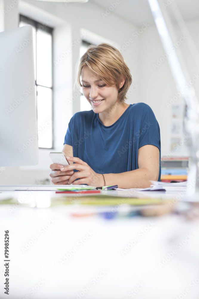 Portrait of smiling young woman using smartphone at her desk in a creative office Portrait of smiling young woman using smartphone at her desk in a creative office