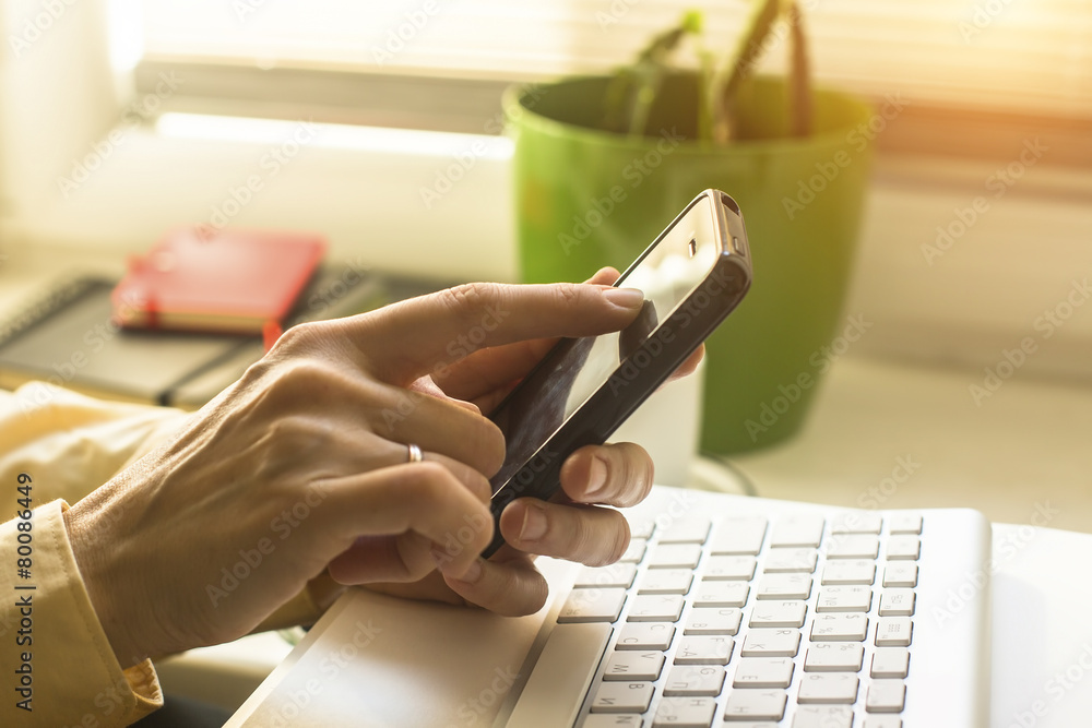 Fototapeta premium Woman using mobile phone, computer keyboard in the background.