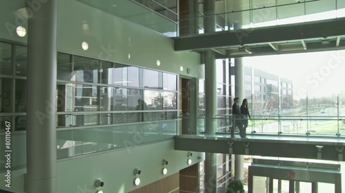 Backlit businessman and woman cross a walkway in a modern building atrium against a backdrop of white windows. Wide shot from same level as people. photo