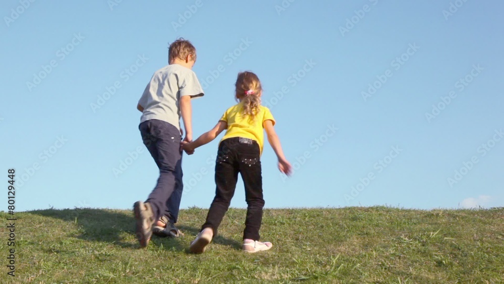 Two kids boy with little girl hold hands and walk away by grass hill at sunny summer day Two kids boy with little girl hold hands and walk away by grass hill at sunny summer day
