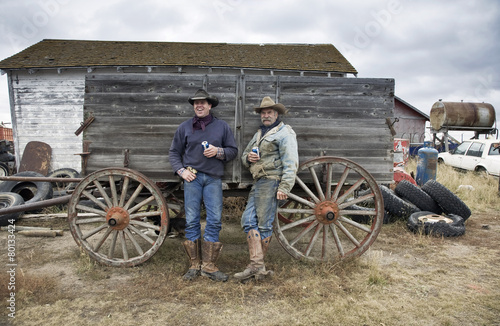 Two men in cowboy hats and cowboy boots leaning against a wooden wagon. photo