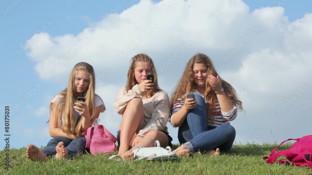 Three girls with bags sit and uses their cell phones on grass hill at sunny summer day Three girls with bags sit and uses their cell phones on grass hill at sunny summer day