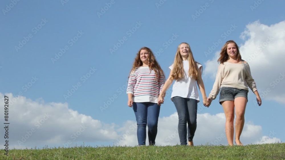Three young girls hold hands and walk closer from behind of grass hill at sunny summer day Three young girls hold hands and walk closer from behind of grass hill at sunny summer day