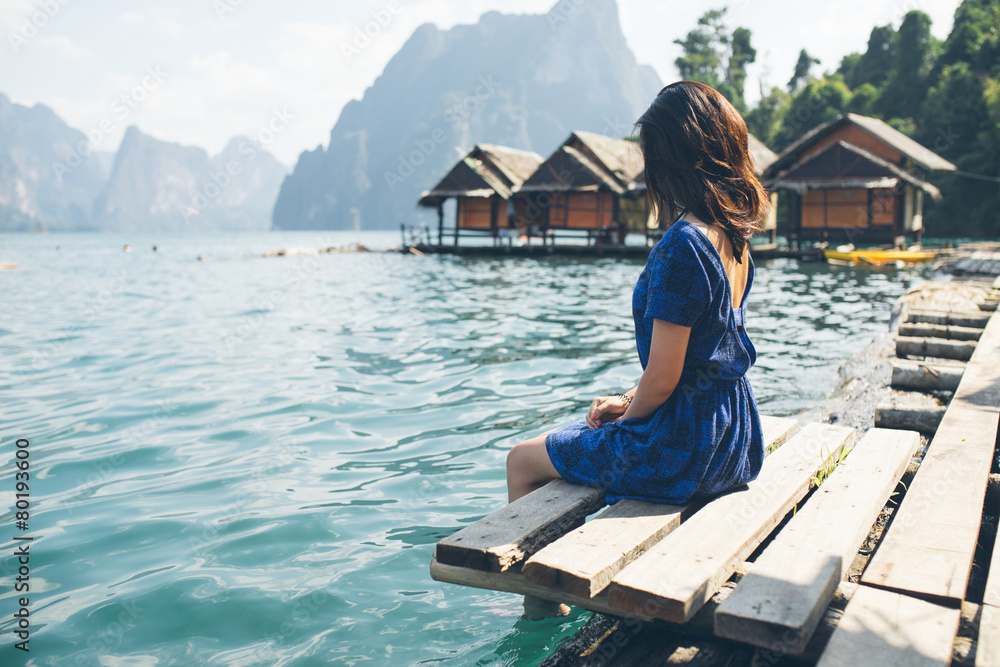 Fototapeta premium Elegant Asian girl wearing blue dress sitting by huge lake surrounded by limestone cliffs in Khao Sok National Park, Phang Nga province, Thailand