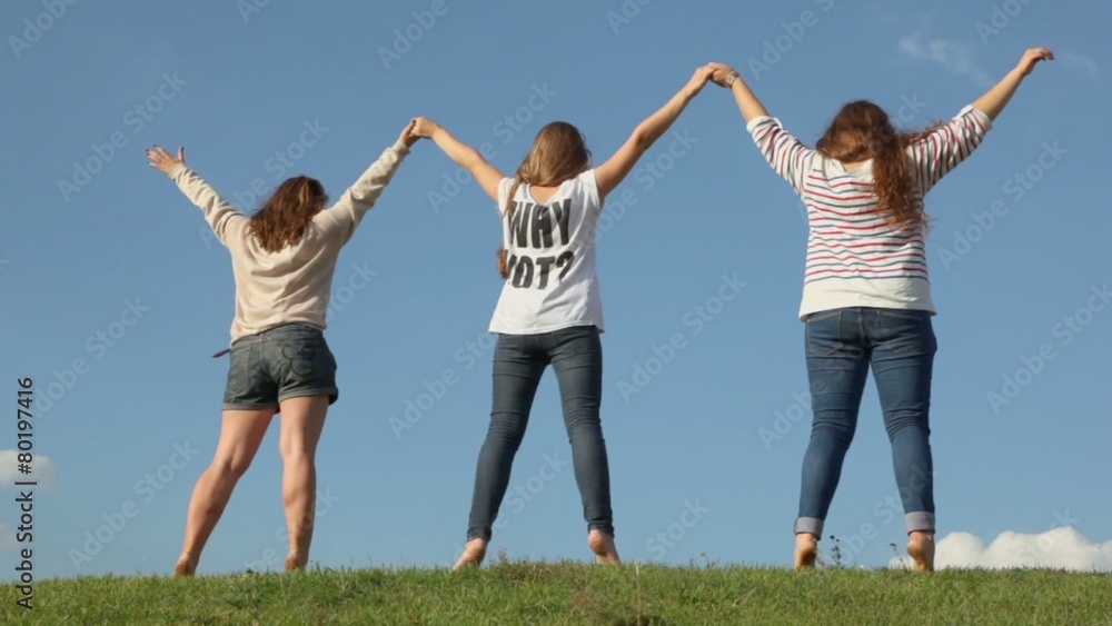 Three young girls hold hands and raise them on grass hill, view from behind at sunny summer day Three young girls hold hands and raise them on grass hill, view from behind at sunny summer day